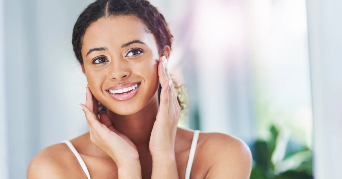 Headshot of a smiling young woman with radiant skin, touching her cheeks, receiving Collagen Stimulating Injections in Coral Springs, FL, in a bright indoor setting.